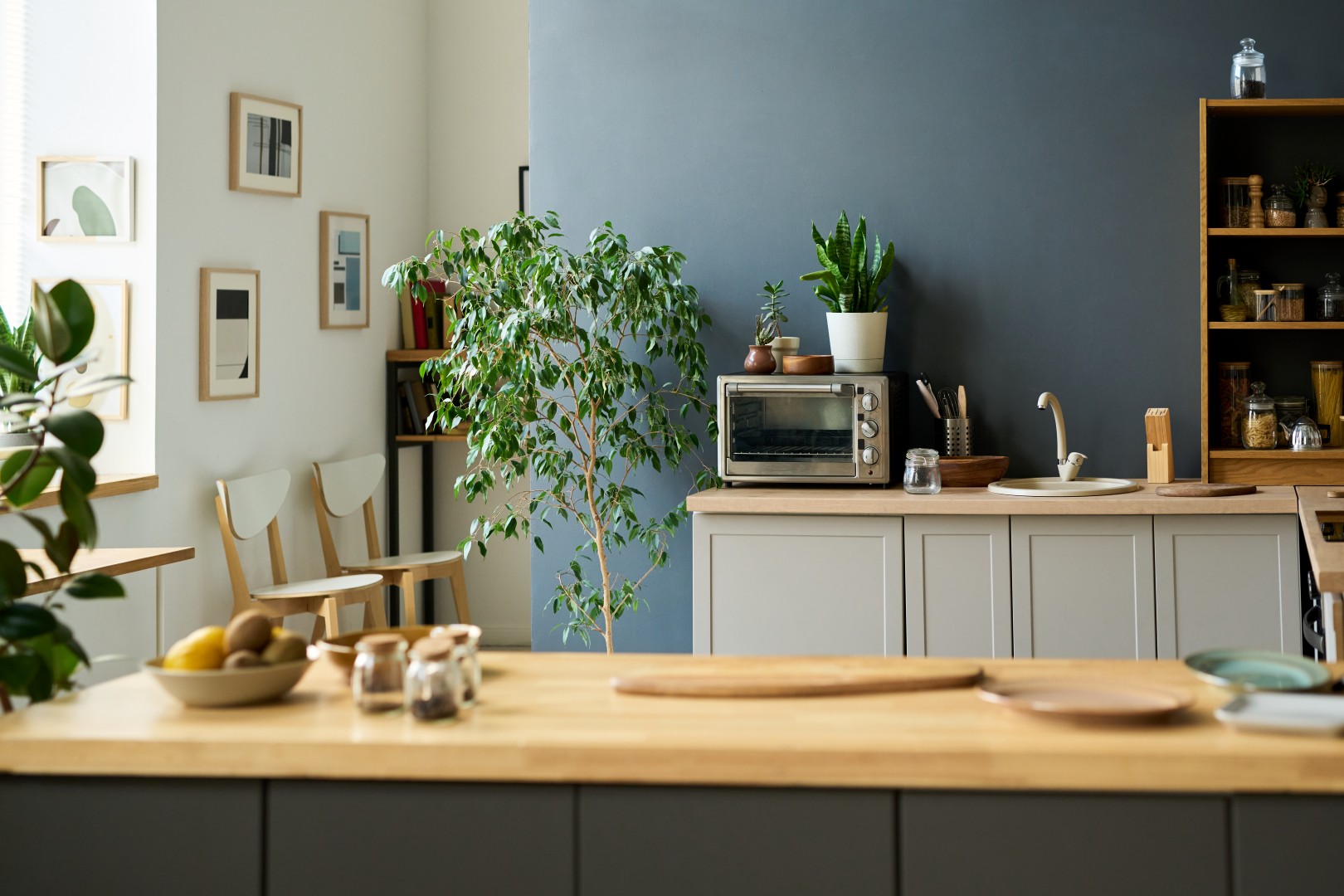Modern kitchen interior featuring wooden countertop, potted plants, microwave oven, kitchen utensils, framed wall art, natural light illuminating clean organized space